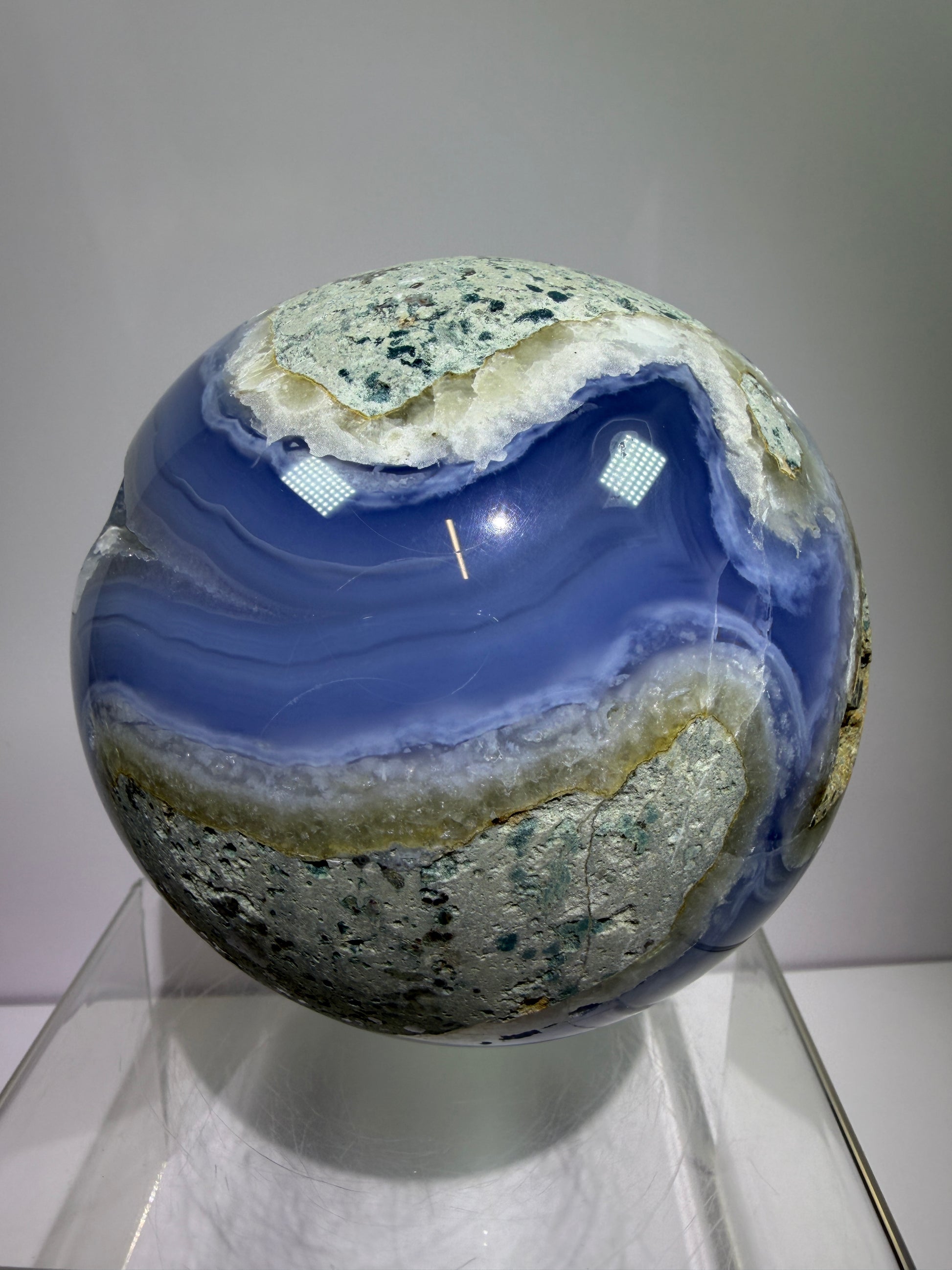 Polished blue lace agate sphere with vivid banded blue and white layers and natural rough matrix on top, displayed on a clear stand against a plain gray background.