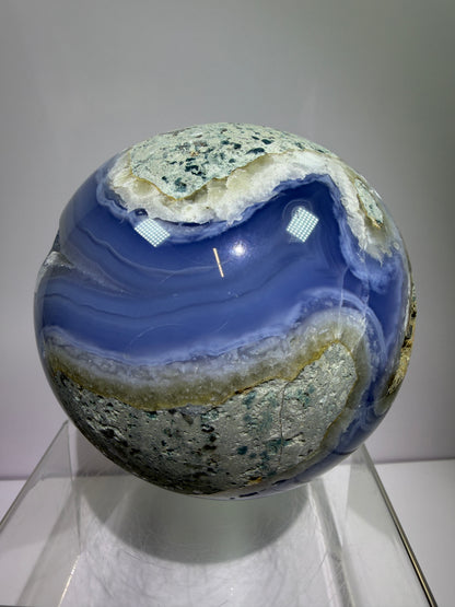 Polished blue lace agate sphere with vivid banded blue and white layers and natural rough matrix on top, displayed on a clear stand against a plain gray background.